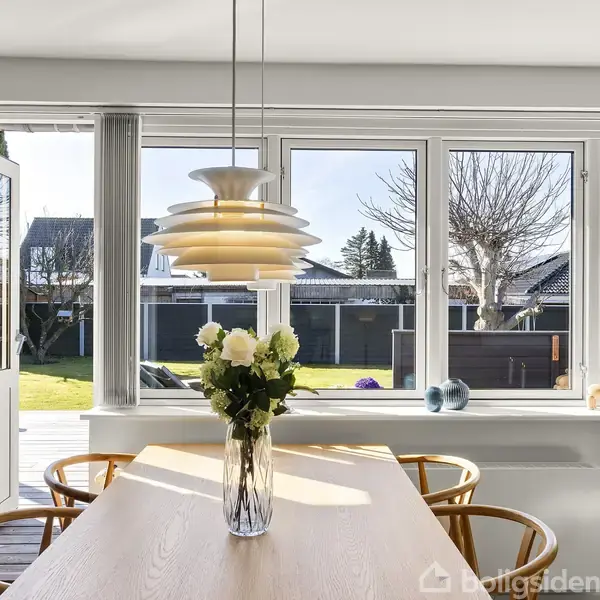 En træspisebord med en vase med hvide blomster står i en lys spisestue. Over bordet hænger en moderne lampe. Gennem vinduerne ses en have med træer og græsplæne.