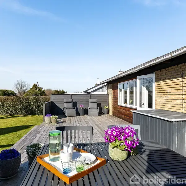 Terrasse med bord, hvorpå der står en bakke med vandkaraffel og glas, omgivet af blomsterkrukker. I baggrunden ses en hæk, græsplæne og en murstensbygning under en blå himmel.