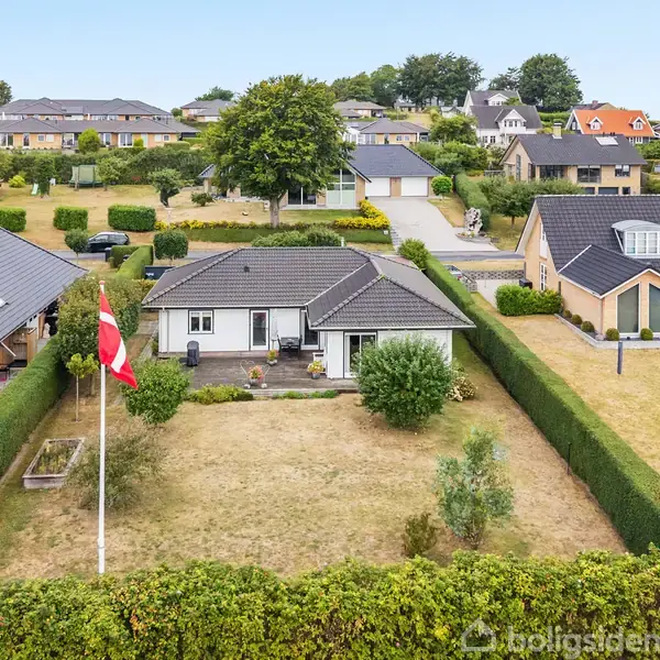 Hus i parcelhuskvarter med sort tag og hvide vægge. Foran ses en velplejet have med hæk og flagstang. Omgivet af lignende huse på en bakketop med grønne omgivelser.
