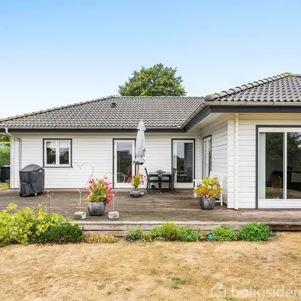 Et hvidt, moderne hus står stille, omgivet af en stor græsplæne og en terrasse med blomsterkrukker og havemøbler.