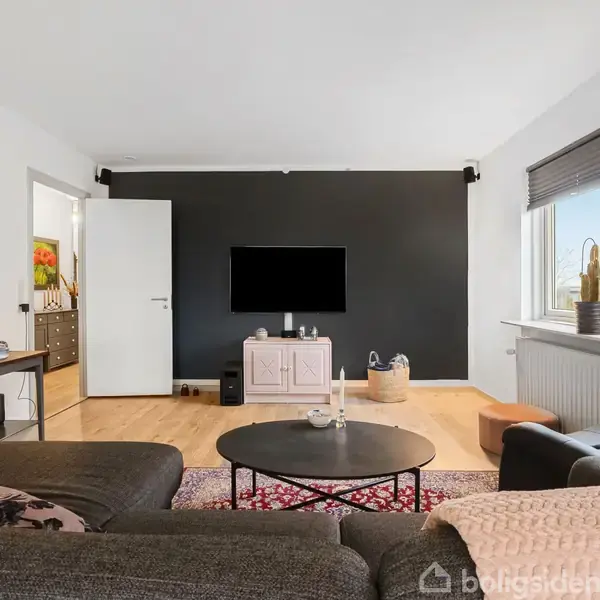 En stue med en grå sofa og rundt sofabord står foran en sort væg med et væghængt TV. En åben dør fører til et tilstødende rum; et vindue med kaktus ved siden lyser rummet op.