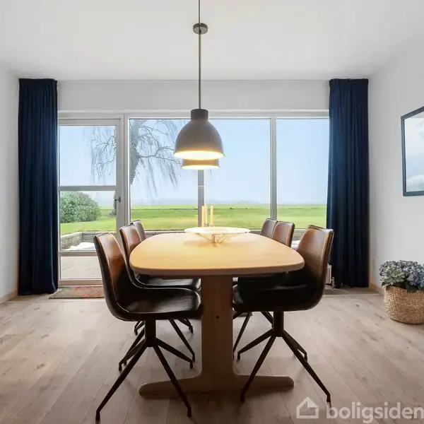 Et træbord omgivet af fire stole står i en spisestue med en stor vinduesfront, der åbner mod en grøn græsplæne udenfor. En lampe hænger over bordet.