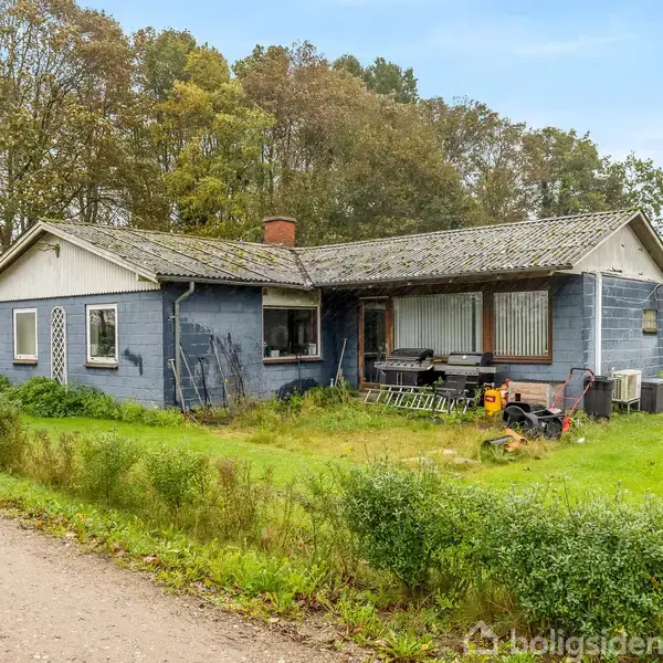 Et gråt hus står i en have omgivet af græs og buske. Der er nogle redskaber og en græsslåmaskine foran huset, og store træer i baggrunden. Vandmærket ignoreres.