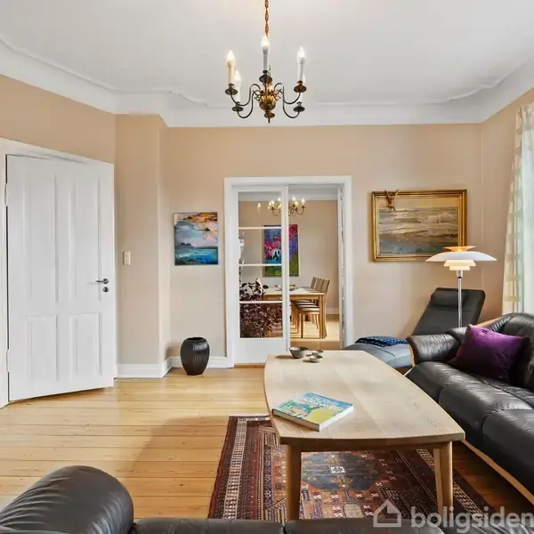 Sort lædersofa står i en stue med lyst trægulv, et træbord foran og en stor lampe til højre. I baggrunden er der en glasdør mod et spiseområde.