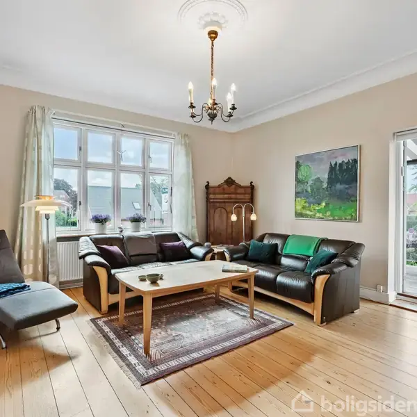 Sofaer i læder står i en stue med parketgulv og lyse vægge. Et sofabord er placeret mellem dem. Store vinduer og en åben dør fører ud til en terrasse.