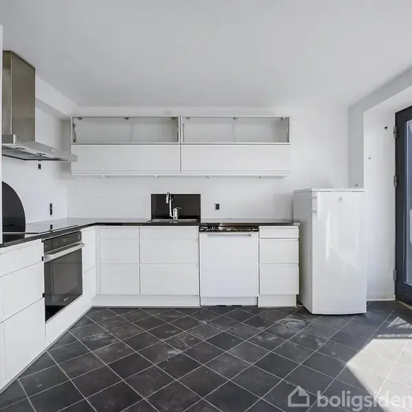 Køkken med hvide skabe og sorte fliser, hvor ovn og køleskab er placeret. En dør med glasparti leder ud til en altan eller terrasse i højre side.