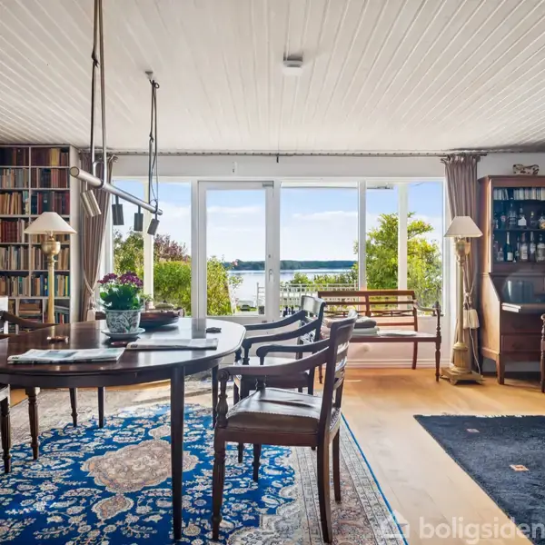 Et spisebord omgivet af stole står på et orientalsk tæppe i et lyst rum med store vinduer, der giver udsigt til en have og vand. Bogreoler dækker væggene.