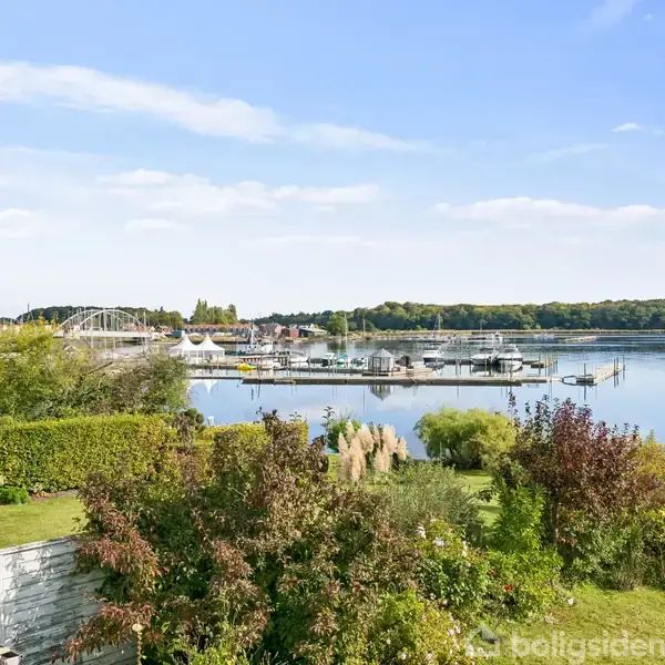 Haven med frodig vegetation ved siden af en sø eller fjord med både og en bro i baggrunden, omgivet af træer og grønne områder under en klar blå himmel.