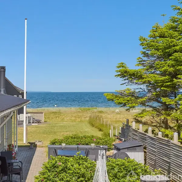 Et sort hus med terrasse står ved siden af en grussti. I baggrunden ses en grøn have, en strand og havet under en klar blå himmel.
