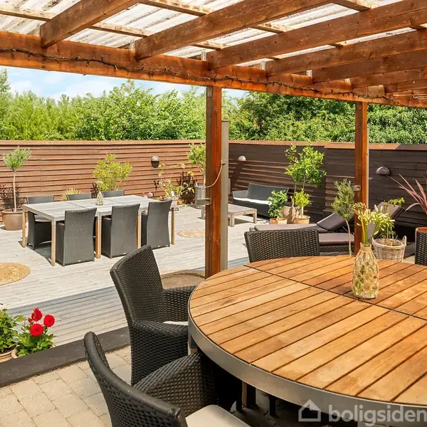 En rund, træbeklædt havebord står under en overdækket terrasse. I baggrunden ses et trædæk med spisebord og stole, omgivet af planter og en grill.