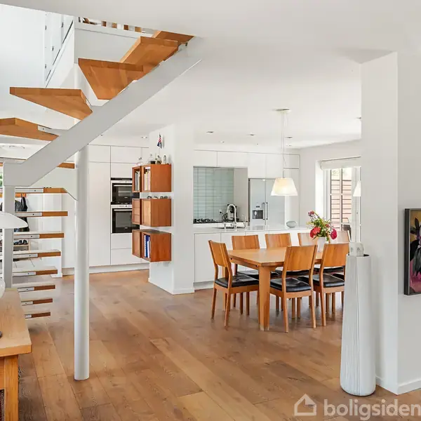 Trappe i træ og metal fører opad i et moderne, lyst køkken-alrum med spisebord og blomster på bordet. Hvide vægge og trægulve. TV på væggen i baggrunden.