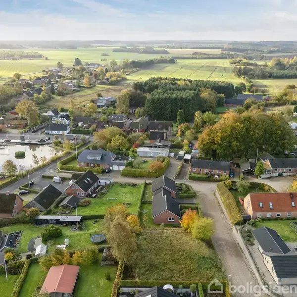 En række huse i en landlig landsby ses fra luften, omgivet af grønne marker og skove. Husene står i rækker langs veje, mens nogle damme er synlige i baggrunden.