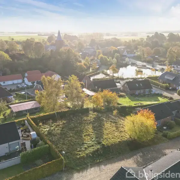 En åben byggegrund ligger omgivet af parcelhuse med haver i et landsbyområde. I baggrunden ses marker og en kirke, omgivet af træer i efterårsfarver.