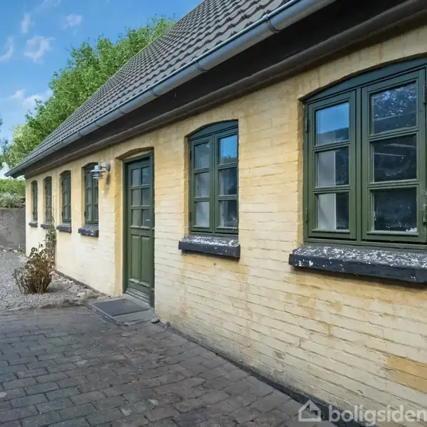 Gul murstensbygning med grønne døre og vinduer står i en gårdhave belagt med sten. Baggrunden har en blå himmel og grønne træer, der giver et landligt indtryk.