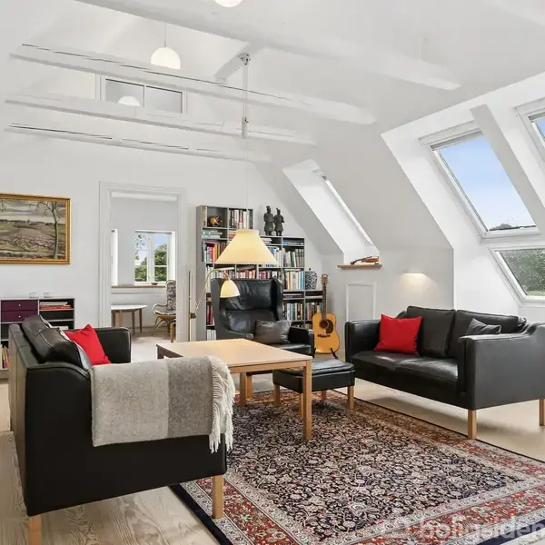 Stue med sorte lædersofaer omkring et træsofabord, en lampe hænger over bordet. Store skråvinduer lader dagslys ind, og en bogreol står i baggrunden ved siden af en guitar.