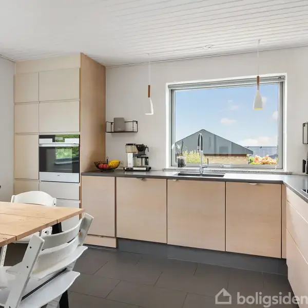 Moderne køkken med lyse træskabe og sorte bordplader. Et stort vindue giver udsigt til en have. Langbord med hvide stole fylder rummet. Lysindfald skaber en lys og rummelig atmosfære.