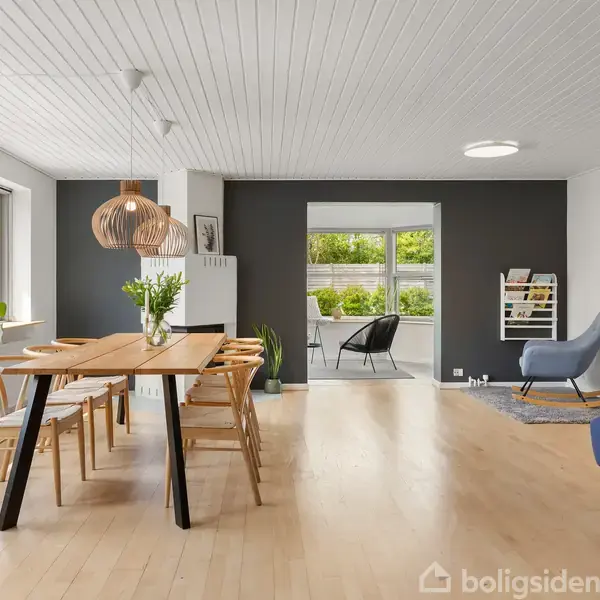 Spisebord med træstole står i en lys stue med lyse trægulve, store vinduer og moderne belysning. I baggrunden er et opholdsområde med stole og grønne planter.