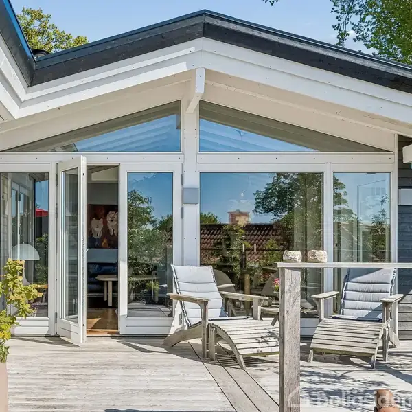 Terrassehus med åbne glasdøre fører til veranda med liggestole. Omgivet af træer og flerfarvede blomster i krukker, skaber en afslappende udendørs stemning.