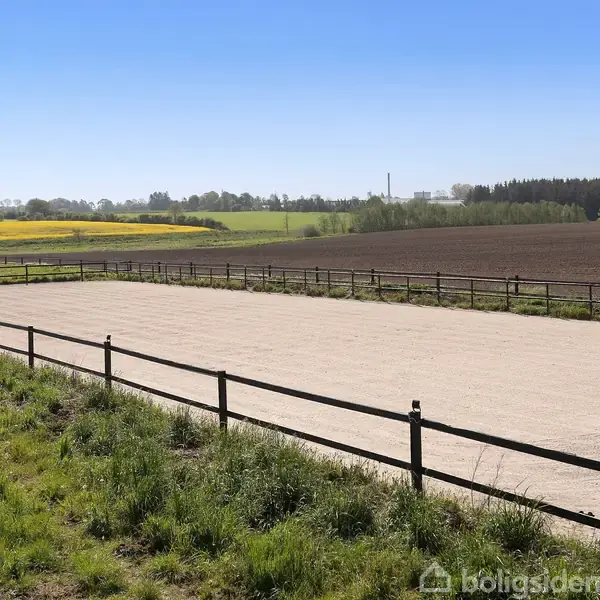 Åben ridebane omgivet af hegn, beliggende i landligt område med marker og skove i baggrunden under en klar blå himmel.