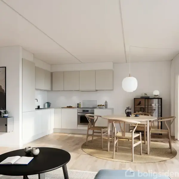 Moderne køkken-alrum med lyse træmøbler, rundt spisebord og stole, og et lille sort sofabord med bøger og lys. Køkkenet har minimalistiske hvide skabe, og en stor hængelampe giver rummet lys.