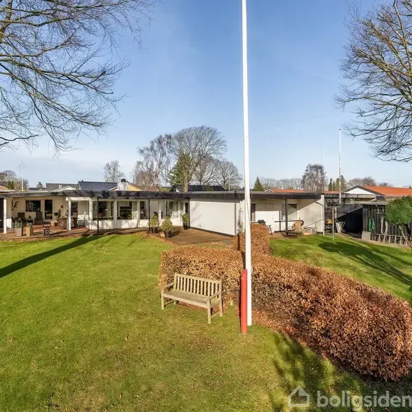 En flagstang står på en velplejet græsplæne med træer og buske omkring. I baggrunden ses en moderne enfamilieshus med en terrasse. En bænk står nær flagstangen.