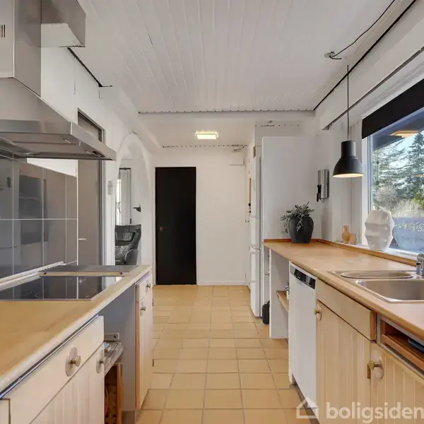 Køkken med træbordplader og rustfrit stål hvidevarer. Lyset træder ind gennem store vinduer med udsigt til træer udenfor. Fliser på gulvet giver en lys og moderne atmosfære.