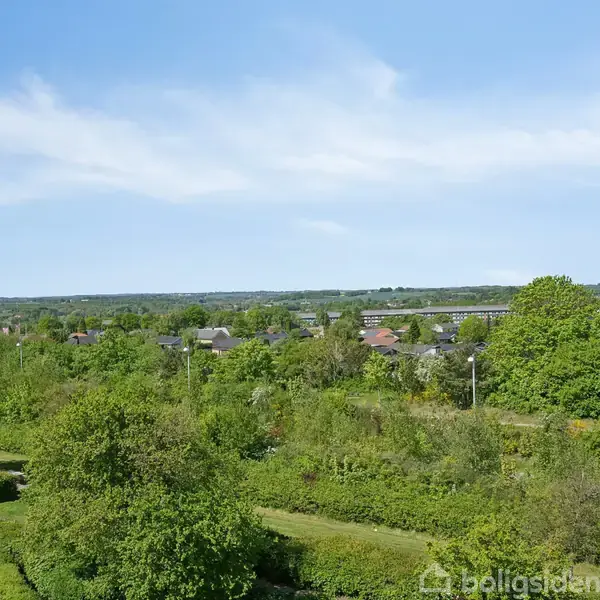 Landskabet strækker sig ud, dækket af frodig, grøn vegetation og spredte træer. Små huse ligger i det fjerne under en klar, blå himmel.
