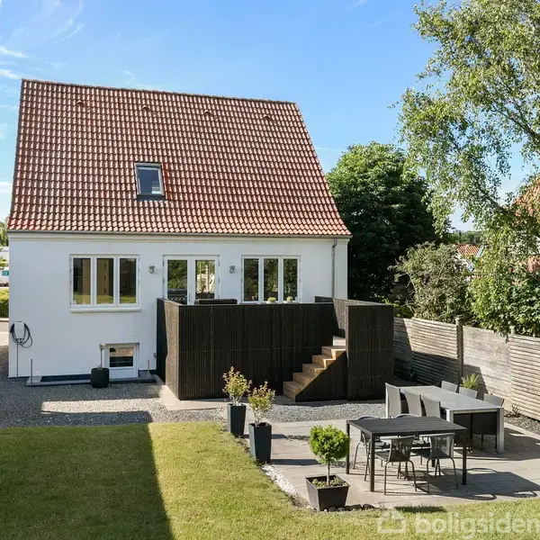 Hus med rødt tegltag står stille i en have med græs og fliseterrasse. Haven er indrammet af et hegn, og der er havemøbler på terrassen under den blå himmel.