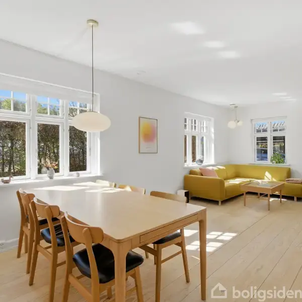 Spisebord med seks stole står i en rummelig, lys stue med parketgulv. Ved siden af ses en grøn sofa under et stort vindue. Væggene er hvide med abstrakt kunst.