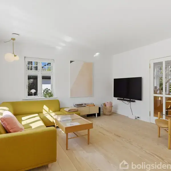 Gul sofa står i stue, belyst af naturligt lys fra vinduer. TV hænger på væggen overfor, ved siden af en bogreol og en dør. Trægulve og hvide vægge fuldender rummet.