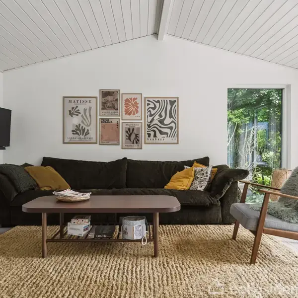 En stor, mørk sofa med farverige puder står i en lys stue. Foran sofaen er et træsofabord på et vævet tæppe. Væggene prydes af kunstplakater, inklusive teksten "Matisse".