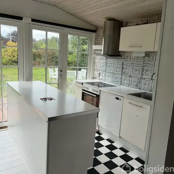 Et moderne køkken med en ø i midten, omgivet af hvide skabe og apparater. Store vinduesdøre åbner ud mod en frodig have med havemøbler. Ingen tekst fundet.