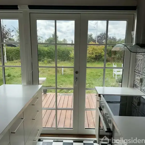 En glasdør står åben ud mod en terrasse med træplanker. Udenfor er der en grøn have med buske og et hvidt havesæt. Himlen er overskyet.
