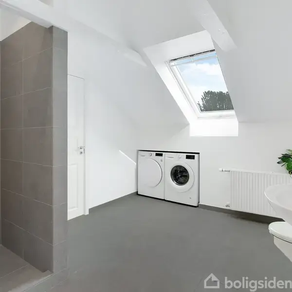 Bruser står på en flisebeklædt væg i et lyst badeværelse med skråloft. Vaskemaskine og tørretumbler er placeret under et ovenlysvindue ved siden af en radiator og en plante.