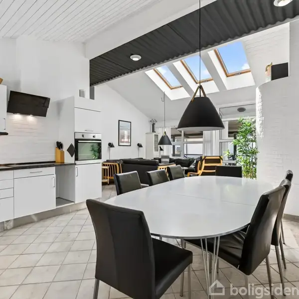 Et moderne køkken med et stort, hvidt spisebord og sorte stole. Ovenlysvinduer lader sollys ind, mens sorte lamper hænger over bordet. Til venstre er der hvide skabe og køkkenudstyr.