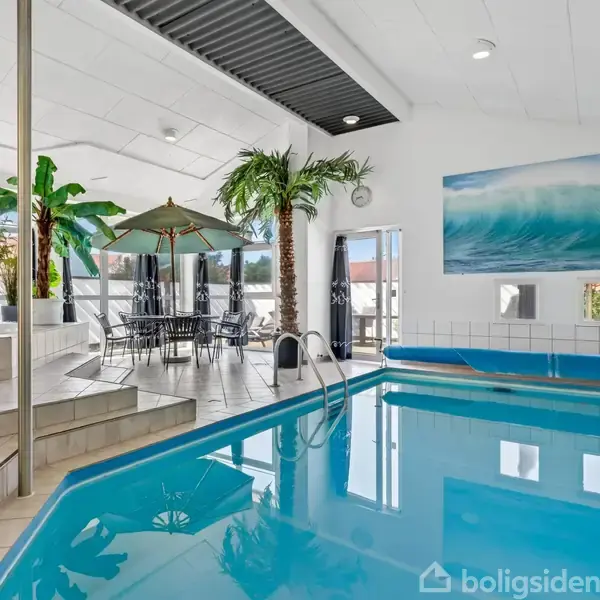 En indendørs swimmingpool er omgivet af tropiske planter og liggestole. Et maleri af en bølge hænger på væggen. I baggrunden ses et bord med parasol ved glasdøre.