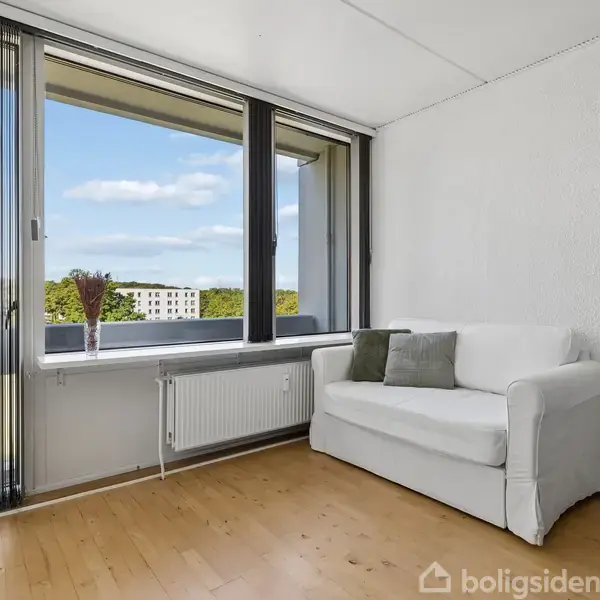 Hvid sofa står foran store vinduer, som lukker lys ind i et værelse med træparket, udsigt over en grøn græsplæne og himmel. En vase med blomster står på vindueskarmen.