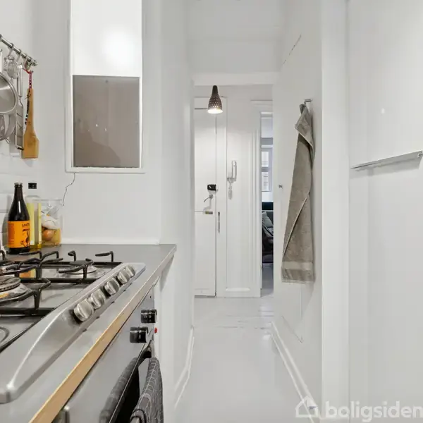 En smal, hvidmalet køkkenkorridor med gaskomfur til venstre. Gryder hænger over komfuret, og en flaske står ved siden af. Højre side har et køleskab.