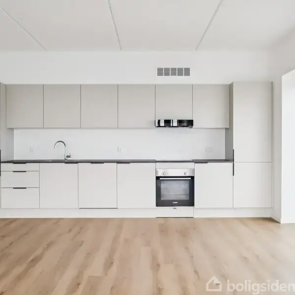 Et moderne køkken med hvide skabe og sølvhåndtag, en indbygget ovn og kogeplade. En stor glasdør til højre fører ud til en altan. Trægulv dækker gulvet.