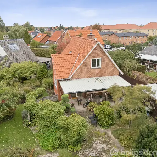 Rødt murstenshus med rødt tegltag står i en frodig have. Huset har en overdækket terrasse med havemøbler. Omkringliggende huse og grønne områder ses i baggrunden.