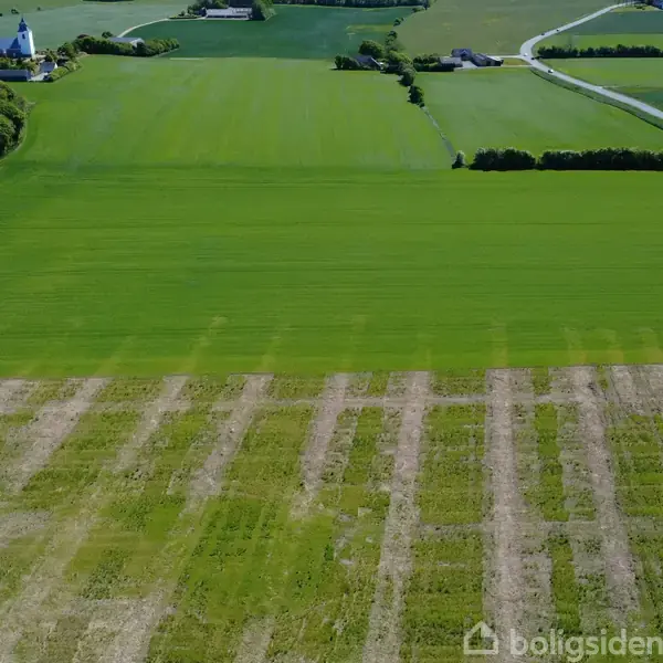 En stor, grøn mark strækker sig under en klar himmel, med en landsbykirke og flere bygninger i baggrunden. Der er også veje og flere marker i det omkringliggende landskab.