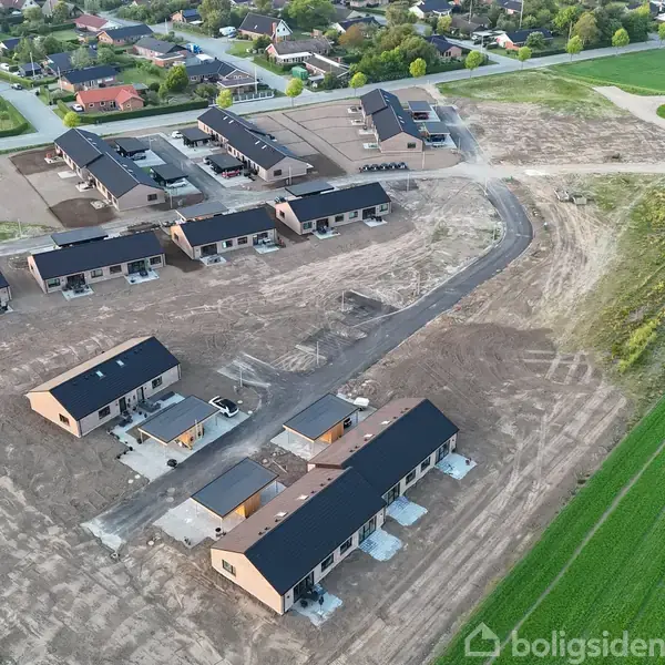 Et nybygget boligområde med rækkehuse, hvor nogle stadig er under opførelse. Husene ligger på en åben mark i et udviklingsområde med nærliggende grønne områder og eksisterende bebyggelse.