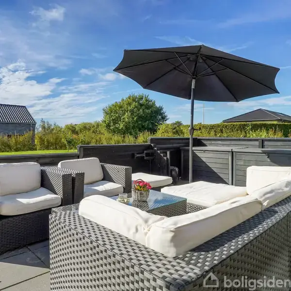 Rattanmøbler med lyse hynder står under en sort parasol. Omkring er en flisebelagt terrasse med grønne buske og en træbygning i baggrunden under en klar blå himmel.