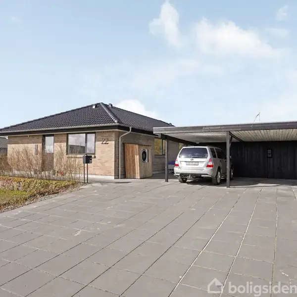 En bil holder parkeret i en carport ved siden af et moderne enfamiliehus i mursten. Området er flisebelagt med en åben, solrig forhave og omgivet af grønne bakker.