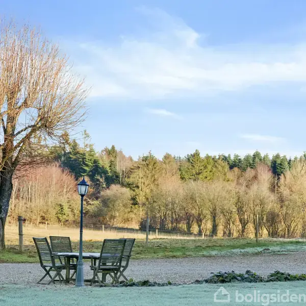 Et bord med fire stole står på en grusvej ved siden af en lygtepæl i en park, omgivet af græsmarker og træer i baggrunden under en blå himmel.