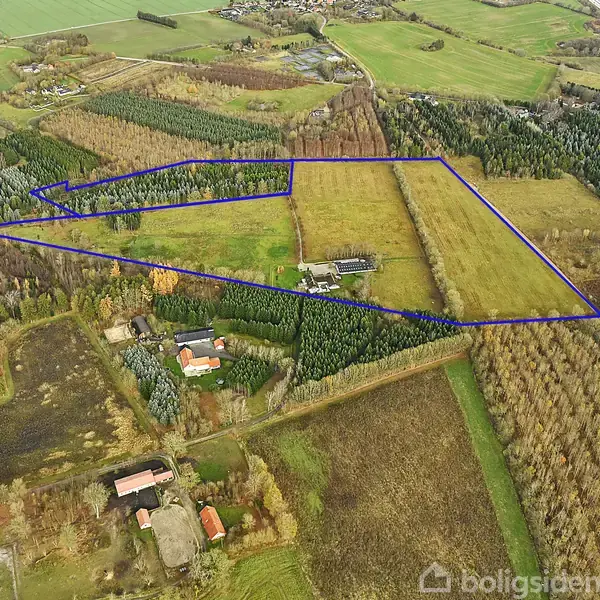 Et stort, afgrænset jordstykke vises fra luften, omgivet af marker, skove og enkelte bygninger i et landligt område med spredt vegetation og veje.