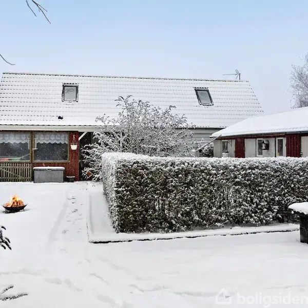 Et sneklædt hus med hæk og træer er omgivet af en gårdhave under en klar, blå himmel. En bålplads er tændt i haven.