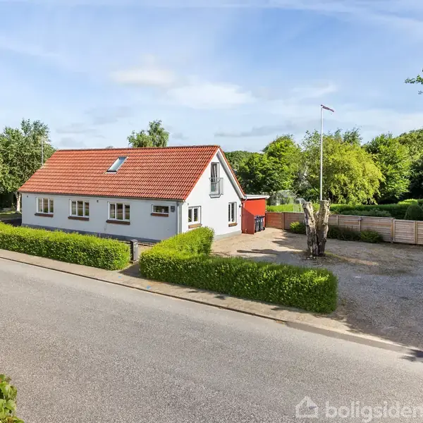 Hvidt hus med rødt tag står i et boligområde. Det er omgivet af en grusparkering og grønne buske. Baggrunden præges af træer og flere huse.