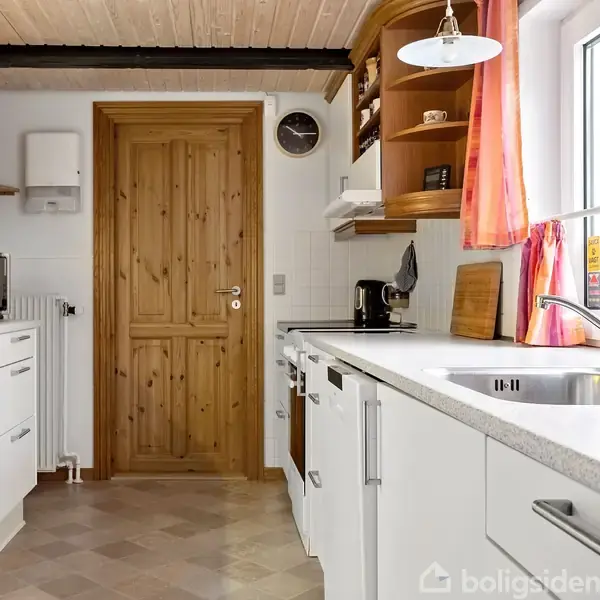 Et lyst køkken har hvide skabe, en opvaskemaskine, og en bordplade med en vask. En mikroovn står til venstre, og et vindue med røde gardiner giver naturligt lys.