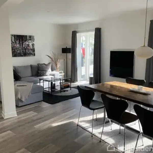 Grå sofa med puder står i en stue ved siden af et sofabord. Rundt spisebord med sorte stole, vinduer og glasdør i baggrunden. Lysindfald gennem gardiner.